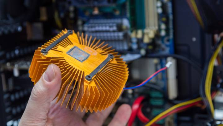Hand holding an orange-plated computer heatsink with PC internals in a blurry background.